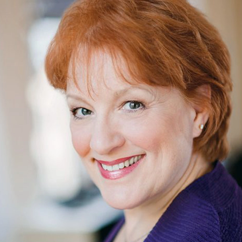 Jeanne Lehman, a woman with short, reddish hair, smiles warmly at the camera. She wears a purple top and small stud earrings. The softly blurred background highlights her friendly expression and bright, light blue eyes.