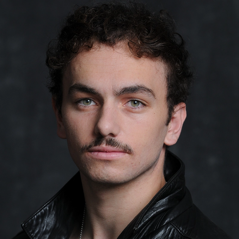 A young man with light skin, curly brown hair, and a thin mustache—styled like Marielle Utayde’s portraits—looks at the camera. He wears a black leather jacket over a dark shirt. The dark, blurred background highlights his serious expression.