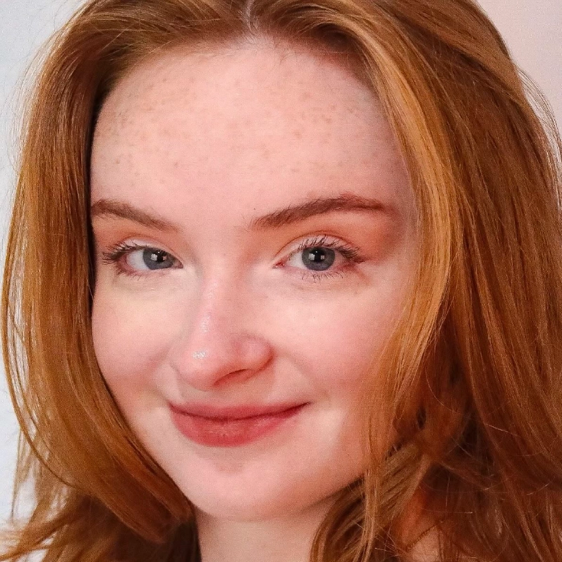 A young person with fair skin, blue eyes, and shoulder-length red hair is smiling softly. Josiah Jennings has light freckles on their face and minimal makeup. The plain, softly lit background draws attention to their friendly expression.