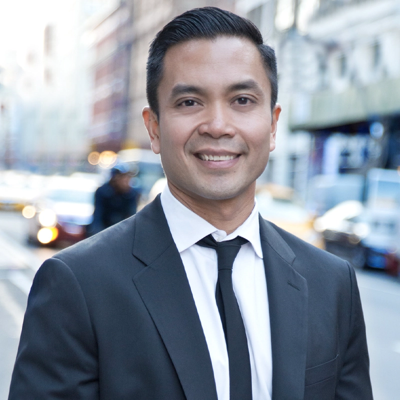 A man in a black suit, white shirt, and black tie stands outdoors on a city street, smiling at the camera. The background is blurred, showing buildings and cars with daylight illuminating the scene.