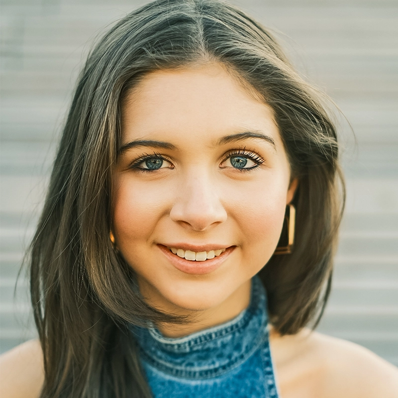 A young woman with long brown hair, blue eyes, and light skin smiles warmly at the camera. Marielle Utayde wears a sleeveless denim top and gold square hoop earrings. Blurred grey steps in the background create a soft, outdoor look.