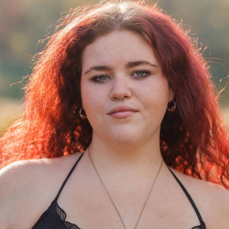 Marielle Utayde, a young woman with long, wavy red hair, stands outdoors. She has light skin, green eyes, and wears a thin black strap top with a nose ring, hoop earrings, and a necklace. The softly blurred background shows greenery and sunlight.