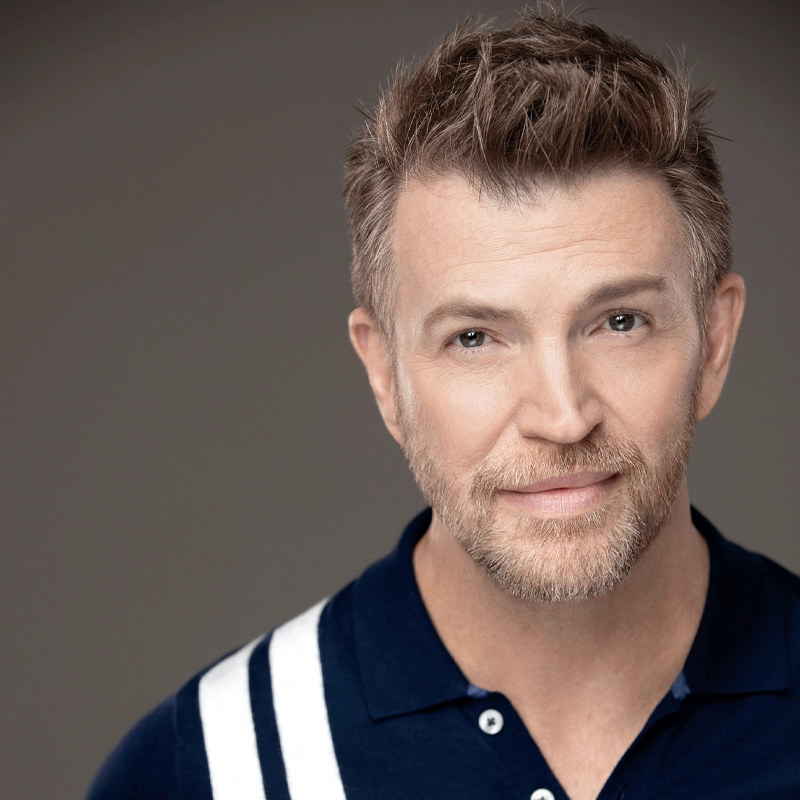 Robert Mannis, a man with short, light brown hair and a trimmed beard, smiles slightly. He wears a navy blue shirt with white stripes on the shoulder against a plain, neutral background. Soft lighting highlights his friendly expression.