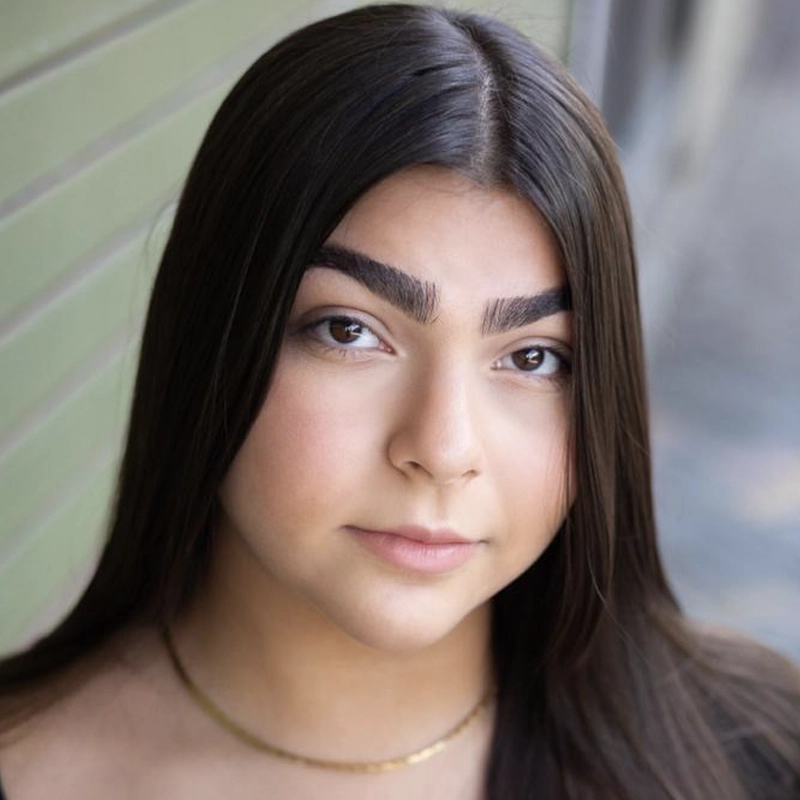 Paola Marcías, a young woman with long, straight dark hair and thick eyebrows, gazes softly at the camera. She wears a simple gold necklace and stands before a green, horizontally lined wall with blurred backgrounds.