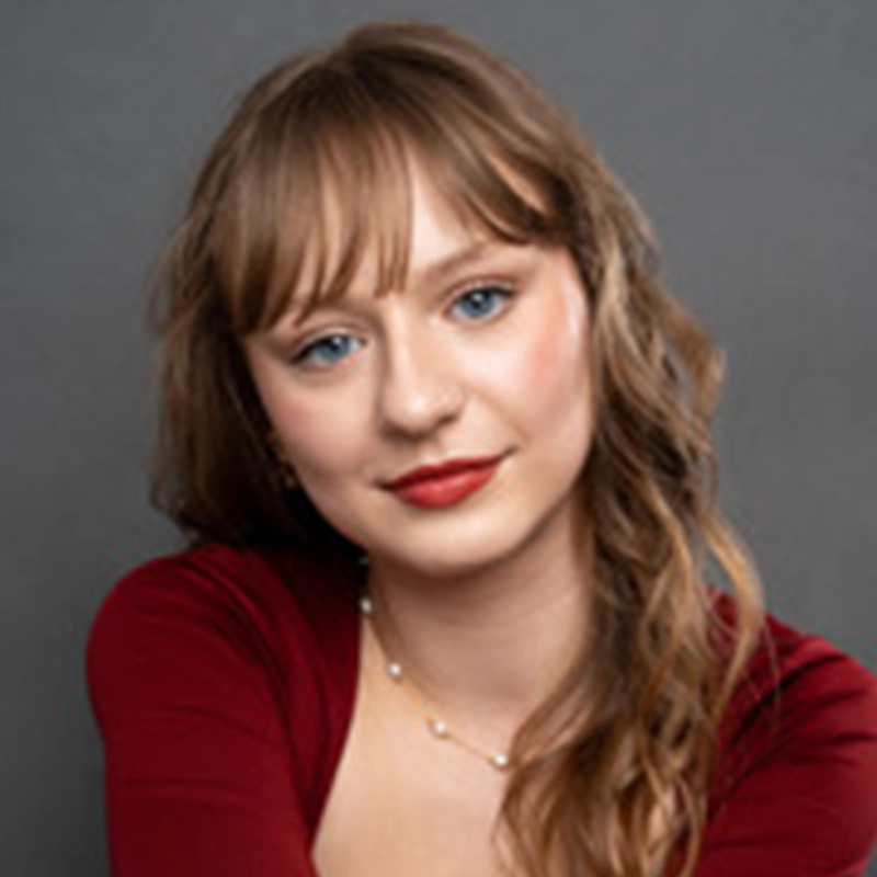 Marguerite A. Boone, a young woman with wavy light brown hair and bangs, smiles softly. She has fair skin, blue eyes, and wears red lipstick. Dressed in a deep red top and delicate gold necklace, she poses against a plain gray background.
