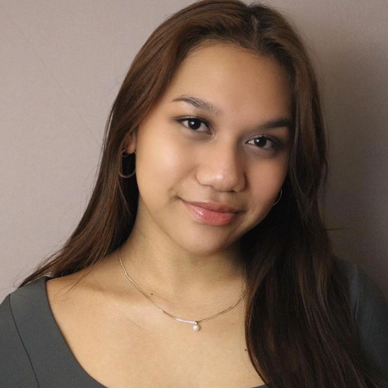 A young woman with long, straight brown hair smiles softly at the camera. Marielle Utayde wears hoop earrings, a delicate gold necklace with a small pendant, and a deep green top. The background is a plain, light beige wall.