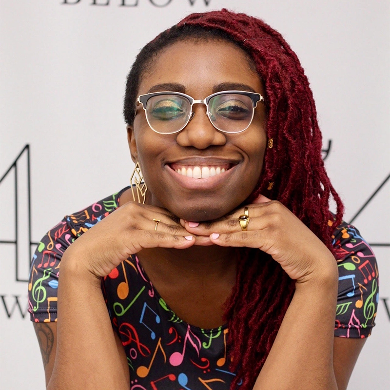 A smiling person with glasses and long red dreadlocks rests their chin on their hands. They wear a colorful shirt with a music note pattern and gold earrings, set against a light background featuring Seth Remington’s partially visible black text and numbers.