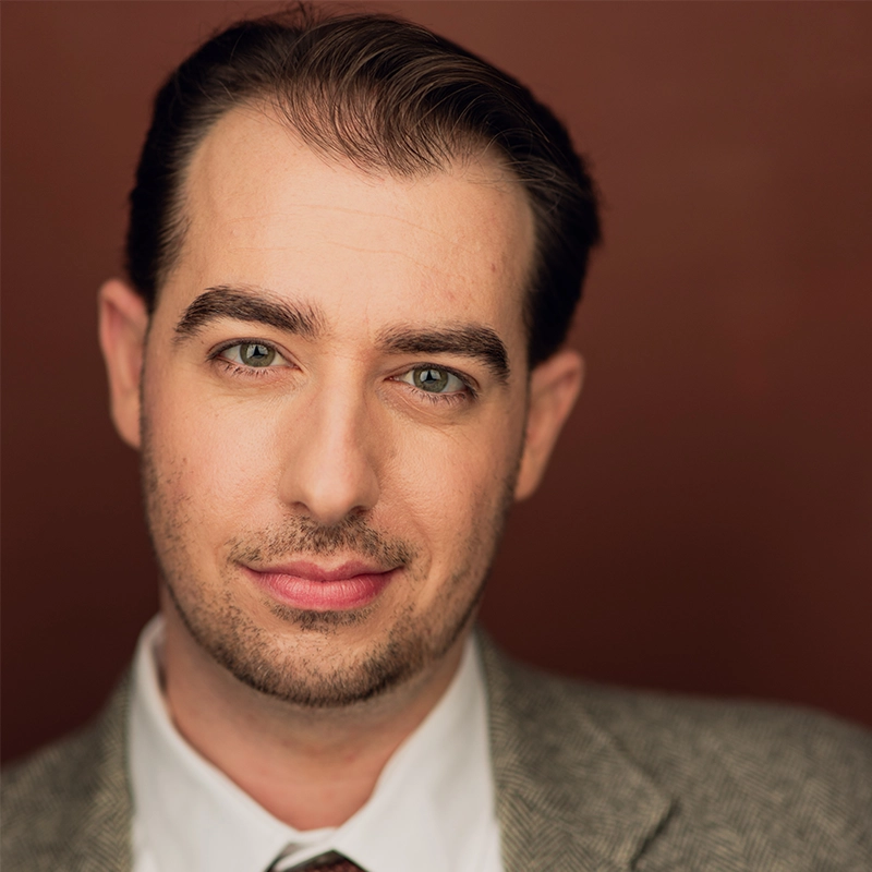 A man with neatly combed dark hair, light skin, and hazel eyes looks directly at the camera. He has trimmed facial hair and wears a white collared shirt, patterned tie, and gray blazer—photographed by Amanda Harris against a warm brown background.