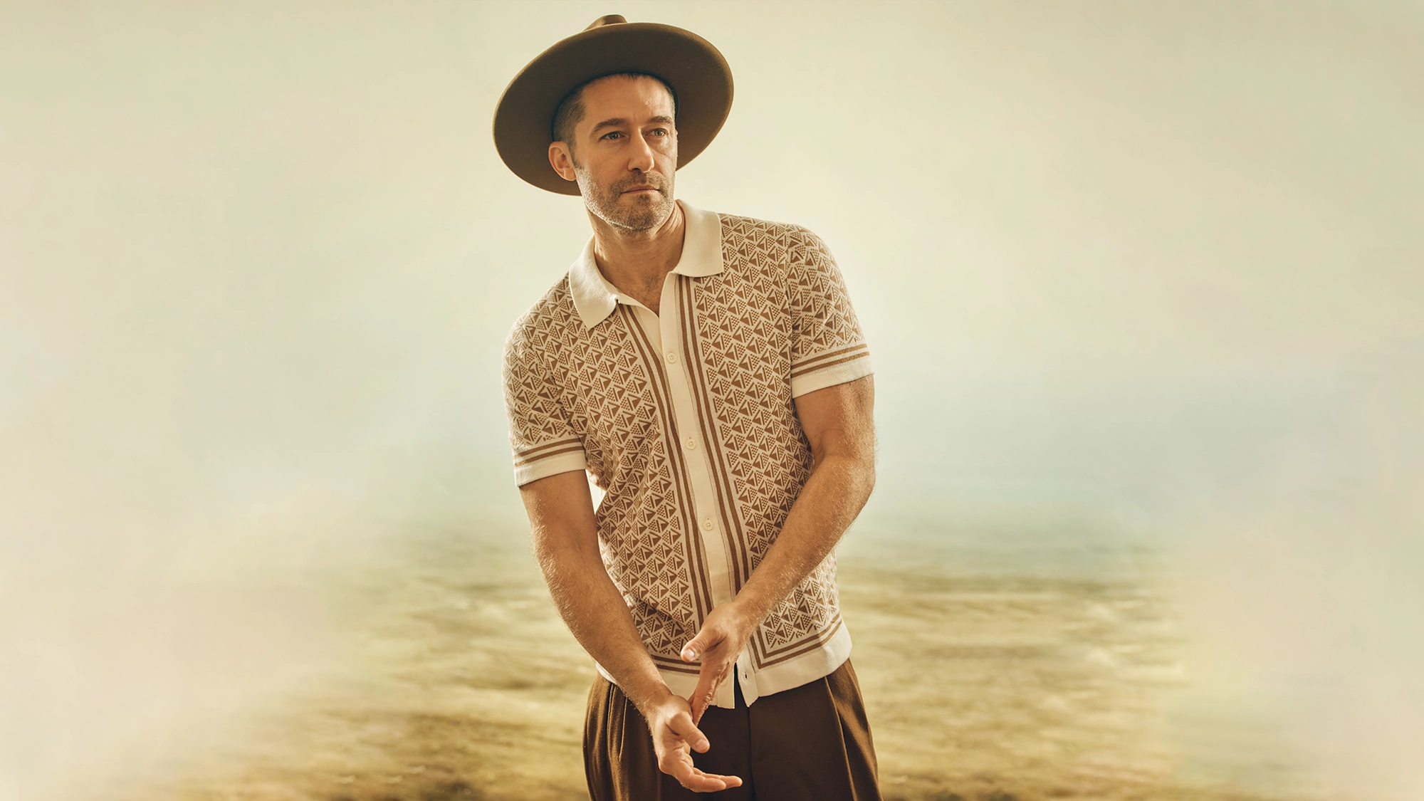 A man wearing a wide-brimmed hat, patterned short-sleeve shirt, and brown pants stands in a sunlit outdoor setting with a hazy background. Channeling Rhythms & Revelations, he gazes forward calmly, hands clasped near his waist.