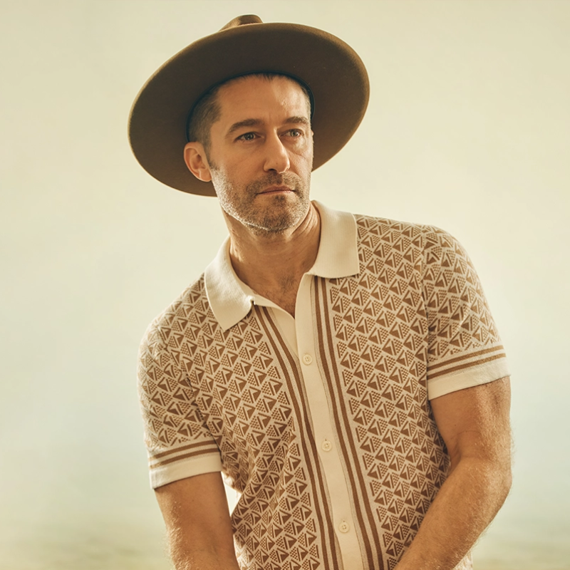 A man in a wide-brimmed hat and patterned short-sleeve shirt poses against a soft, beige background. He has a trimmed beard, looks off to the side thoughtfully, and stands in relaxed lighting, creating a warm, vintage atmosphere.
