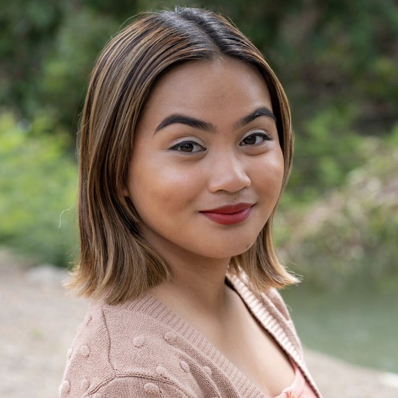 Ella, a young woman with short, light brown hair and smooth skin, smiles slightly at the camera. She wears a beige, textured sweater and red lipstick. The background is outdoors in Venice, with soft green foliage and a blurred natural setting.