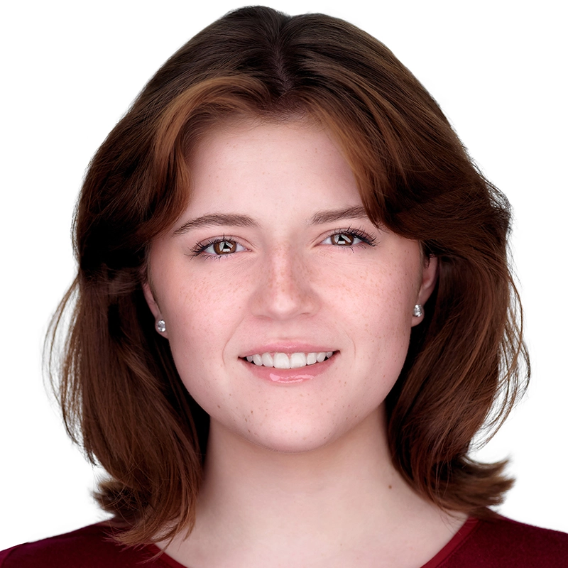 A young woman with shoulder-length brown hair smiles gently at the camera. She has fair skin with freckles, light pink lips, and is wearing small stud earrings. She is dressed in a maroon top against a plain white background, photographed by Josiah Jennings.