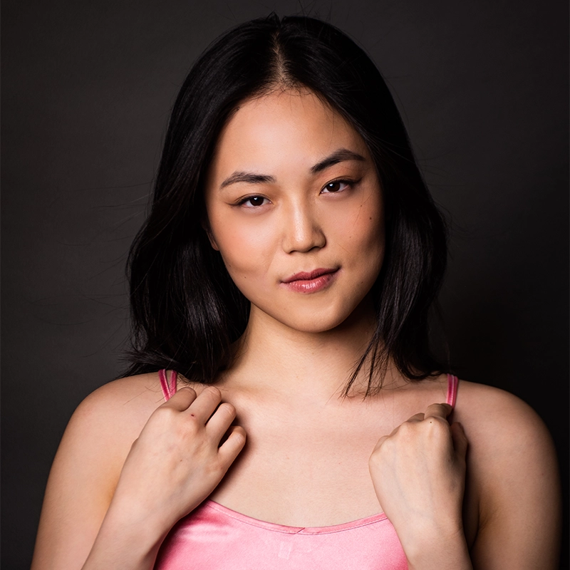 A young woman, Olivia Luzquinos, with straight black hair stands against a dark background, wearing a pink sleeveless top. She looks directly at the camera with a slight smile, holding both straps of her top as light highlights her features.