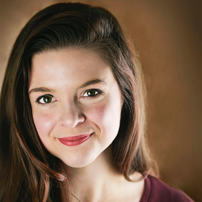 A young woman with long brown hair, Adriana Ellis, smiles softly at the camera. She has light skin, brown eyes, and wears a berry-colored top against a warm, blurred brown background that draws focus to her brightly lit and expressive face.
