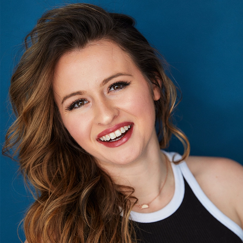 Amanda Harris, a young woman with long, wavy brown hair, smiles brightly in front of a solid blue background. She wears a black and white sleeveless top, a delicate necklace, bold mascara, and pink lipstick as she looks directly at the camera.