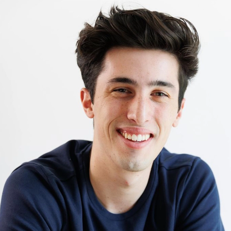 A young man with short, dark, tousled hair smiles at the camera. He has fair skin, dark eyebrows, and is wearing a navy blue shirt. The soft lighting and plain white background create a look Phoebe Eskovitz might capture in her portraits.