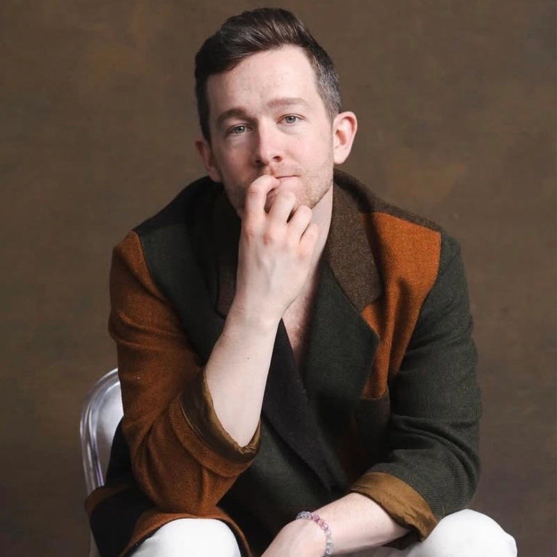 Brady Miller, a man with short brown hair and blue eyes, sits on a chair in a thoughtful pose. He wears a color-blocked brown and green jacket over a dark shirt and white pants, set against a neutral brown backdrop.
