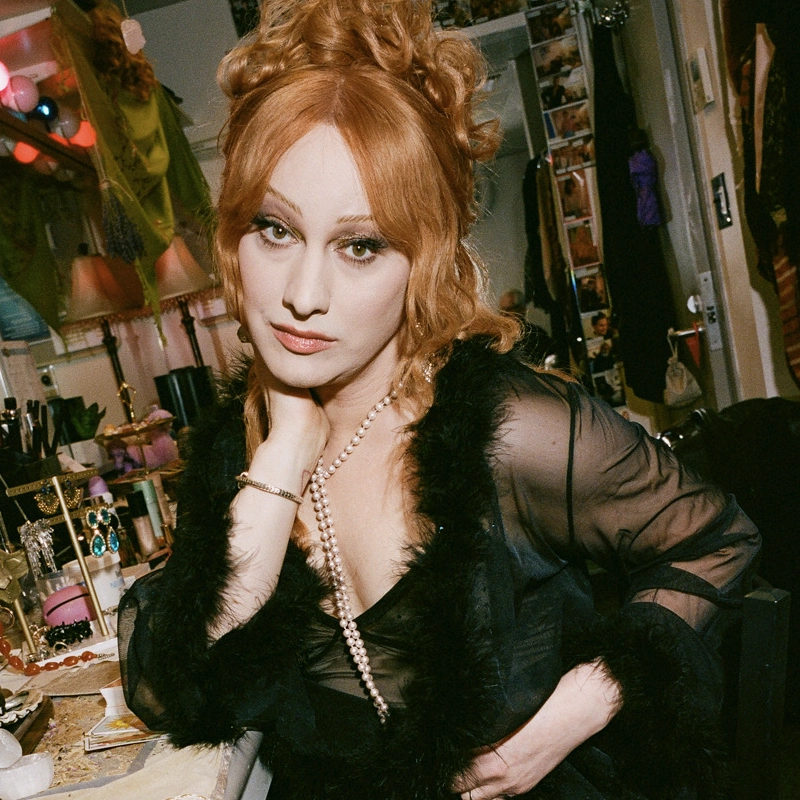 A woman with long red hair styled in loose curls, reminiscent of Jinkx Monsoon, wears a sheer black robe with feather trim and a long pearl necklace. She sits at a cluttered vanity, gazing at the camera with a relaxed, confident expression.