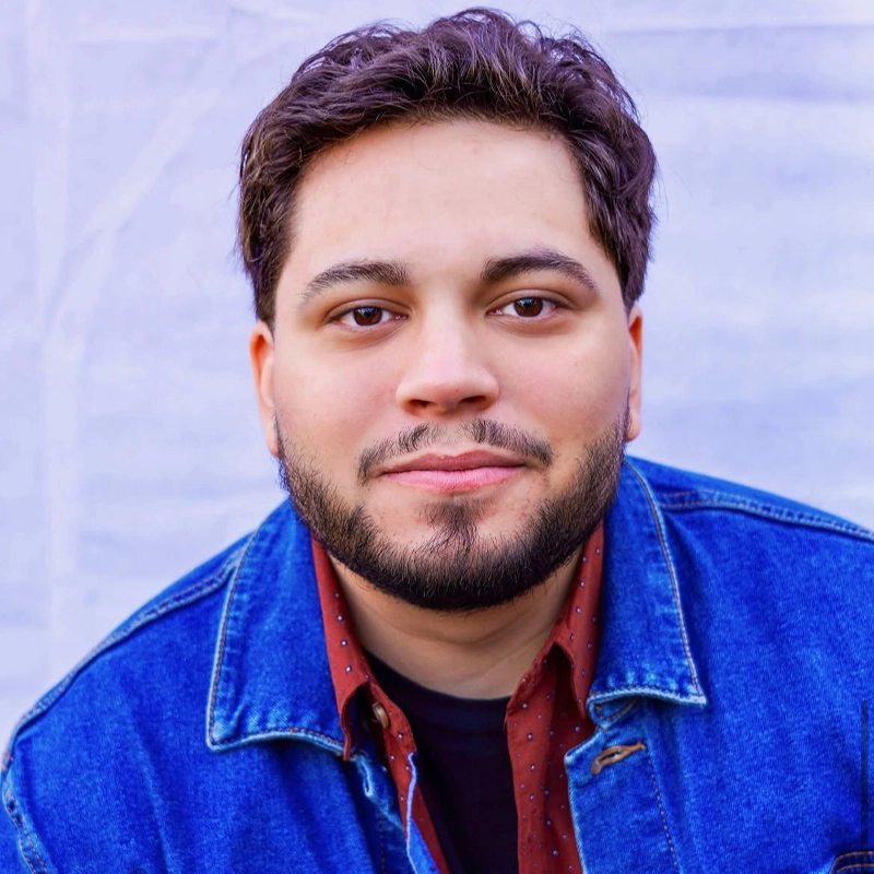 A young man, Joseph Morell, with short, dark, wavy hair and a trimmed beard smiles softly at the camera. He wears a blue denim jacket over a maroon shirt with small white dots. The background is a plain, light purple color.