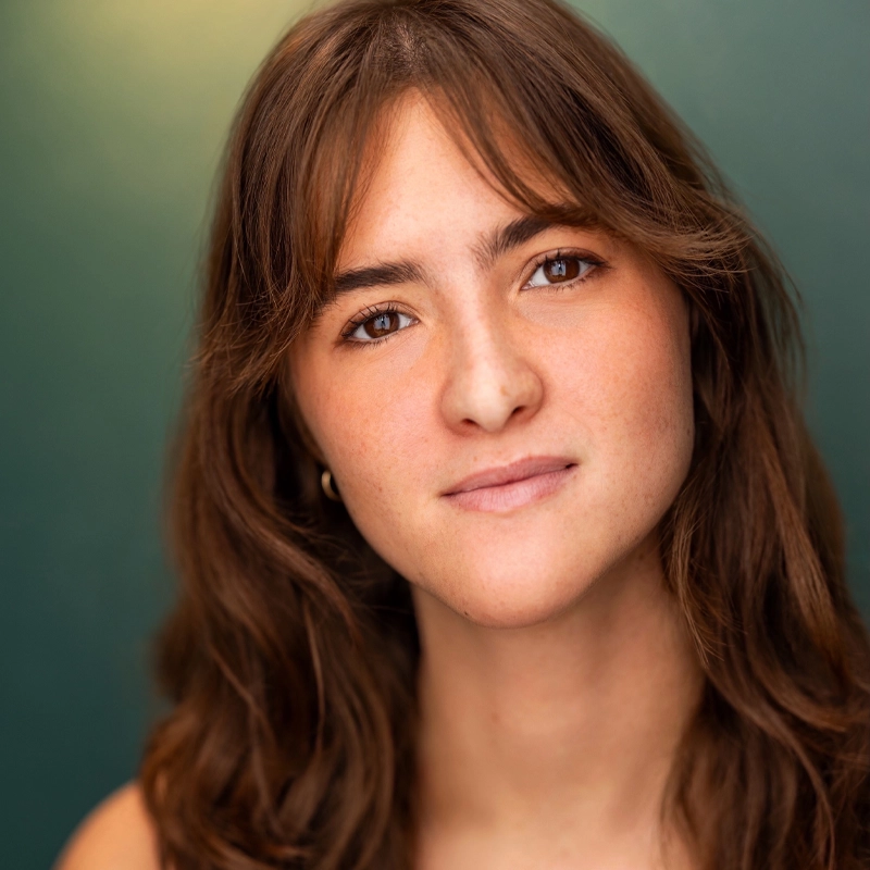 Sophia Muharram, a young woman with wavy brown hair and brown eyes, looks directly at the camera with a neutral, gentle expression. She is wearing a simple top and small hoop earrings against a smooth teal and green gradient background.