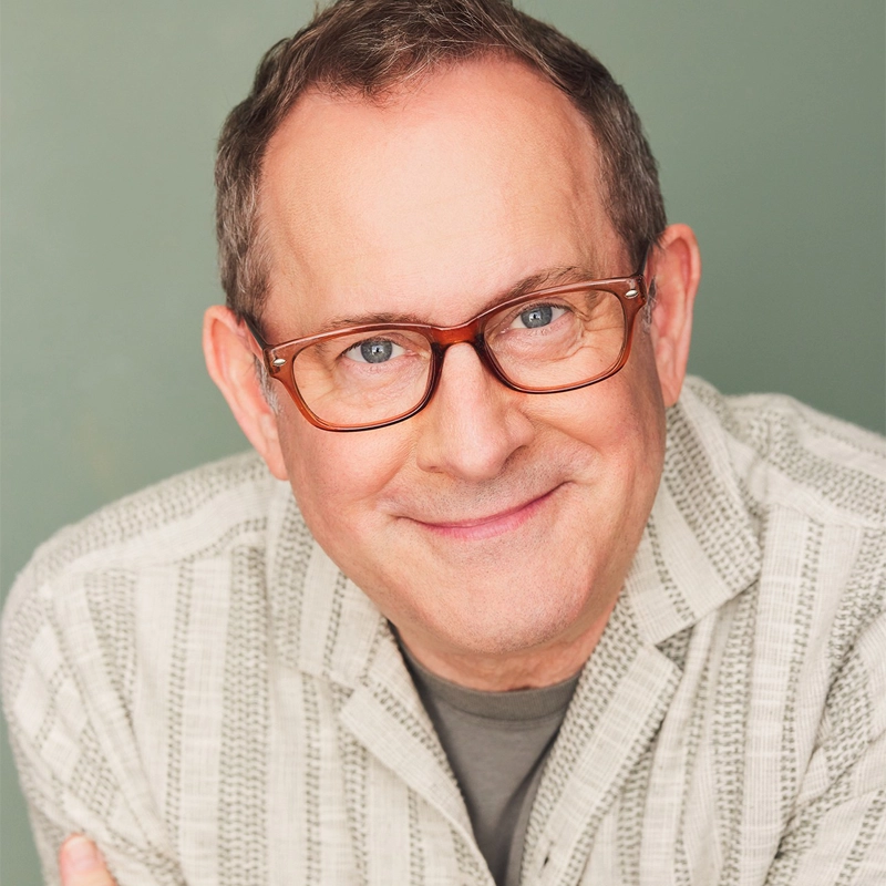 A middle-aged man with short brown hair, wearing brown glasses and a light-colored, patterned button-up shirt, Joel Newsome smiles warmly at the camera against a plain, muted green background.