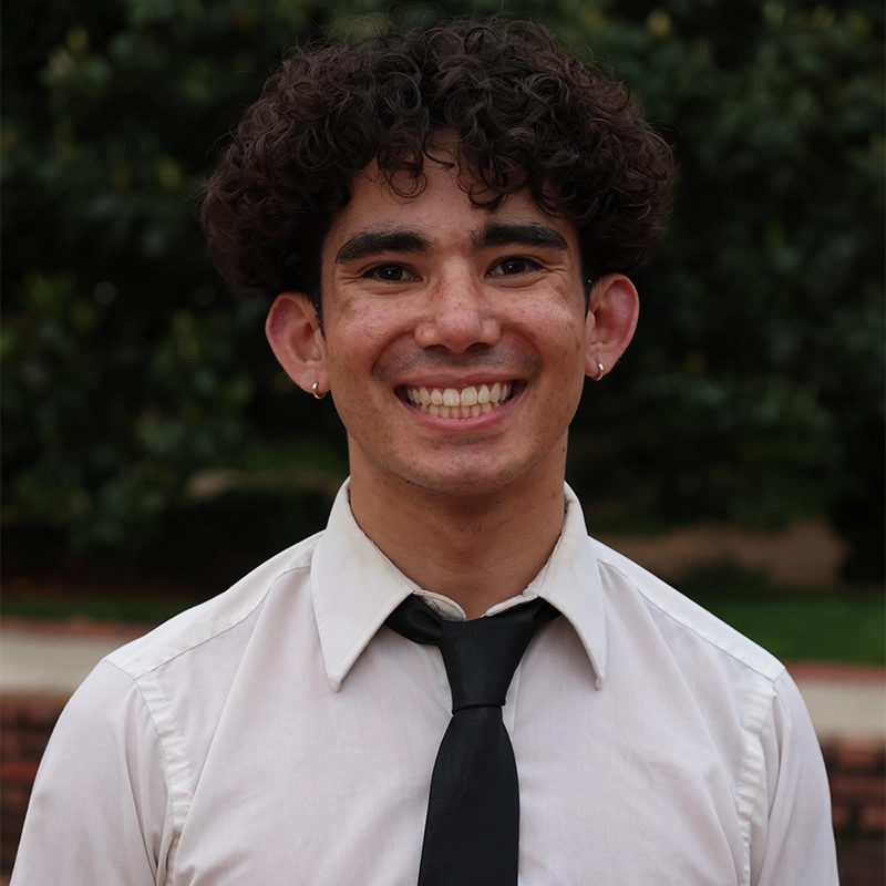A young man with curly dark hair, Nick Benefield, smiles widely at the camera. He wears a light-colored shirt and black tie, with small hoop earrings in both ears. The background is outdoors with green foliage and blurred trees.