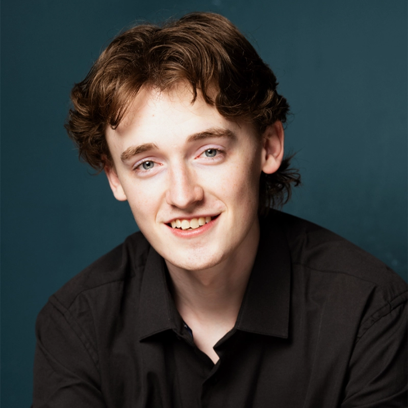 A young man, Seth Remington, with light skin, wavy brown hair, and blue eyes smiles at the camera. He wears a black button-up shirt and poses against a solid dark teal background, softly and evenly lit.
