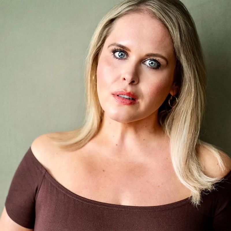 A woman with straight, shoulder-length blonde hair and blue eyes wears a brown off-the-shoulder top. She faces the camera with a neutral expression against a plain, muted green background.