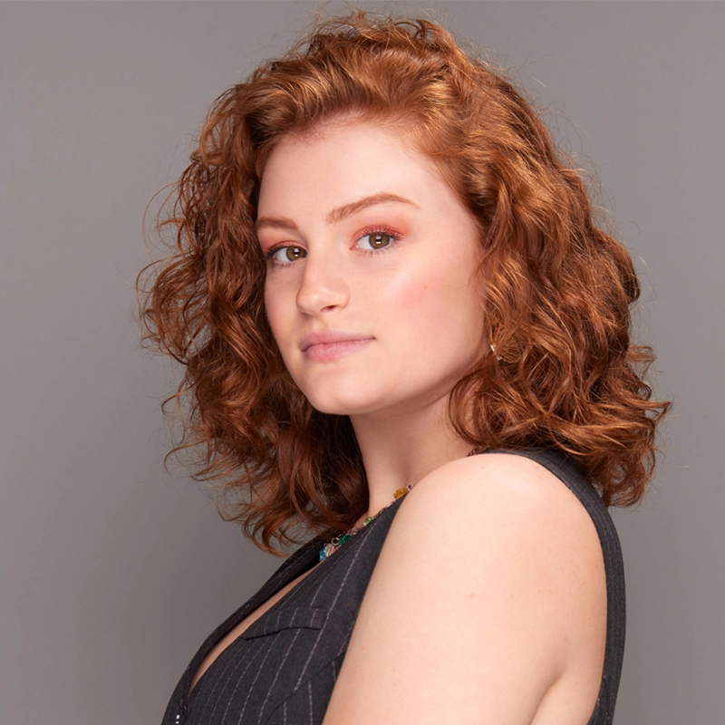 A young woman with fair skin and curly, shoulder-length red hair looks confidently at the camera. She wears a sleeveless dark gray top with subtle stripes, gold hoop earrings, and subtle makeup. The background is a plain, light gray—classic Quincy Lawson style.