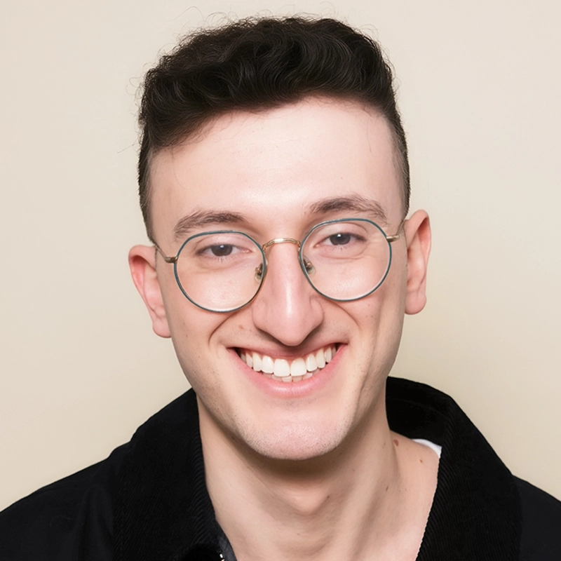Smiling young adult with short, dark, wavy hair and large round glasses, wearing a black jacket over a white shirt, poses against a plain light beige background.