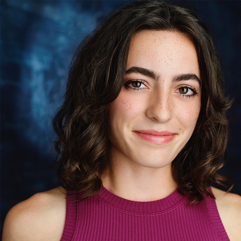 A young person with shoulder-length curly brown hair, arched eyebrows, and light freckles smiles slightly. Olivia Luzquinos wears a sleeveless, ribbed magenta top. The background is a softly blurred mix of dark and light blue tones.