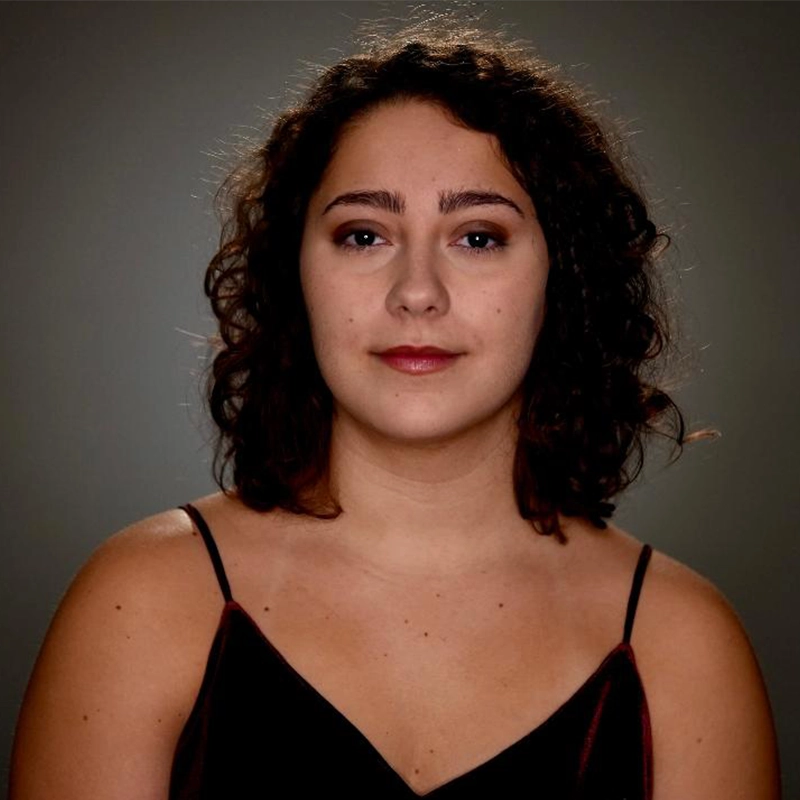 A young woman with curly brown hair and light skin faces the camera, wearing a sleeveless dark top. Olivia Luzquinos has natural makeup with subtle eye shadow and berry lipstick. The background is plain and softly lit, with a slight vignette effect.