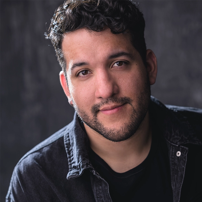 Sergio Guerra, a man with short, curly dark hair and a trimmed beard, smiles slightly. He wears a black t-shirt and a dark, open denim jacket. The background is dark and blurred, highlighting his face with soft, even lighting.