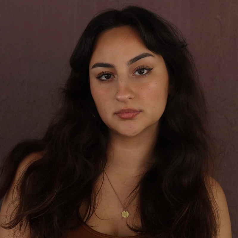 Emma Wallace, a young woman with long, wavy dark hair and arched eyebrows, looks directly at the camera with a neutral expression. She wears a brown tank top and a round gold necklace against a plain, muted brown wall.