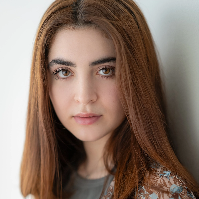A young woman with long, straight auburn hair and light skin gazes softly at the camera. She has full eyebrows, hazel eyes, and wears a patterned top by Sergio Guerra, standing against a plain, light-colored background.