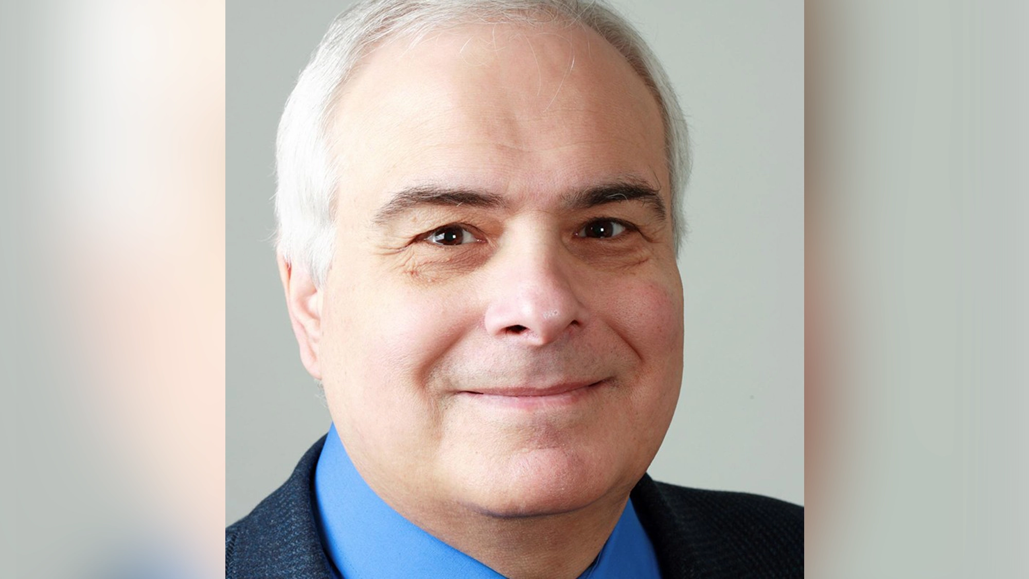 A middle-aged man with short gray hair and fair skin is smiling slightly. He is wearing a blue dress shirt, blue tie, and dark blazer. This close-up headshot of Peter Filichia features a plain background that reflects his Broadway expertise.