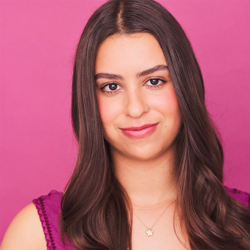 Phoebe Eskovitz, a young woman with long, wavy brown hair, smiles softly in front of a bright pink background. She wears a sleeveless purple top and a gold necklace with a star pendant, her natural makeup featuring rosy cheeks and pink lipstick.