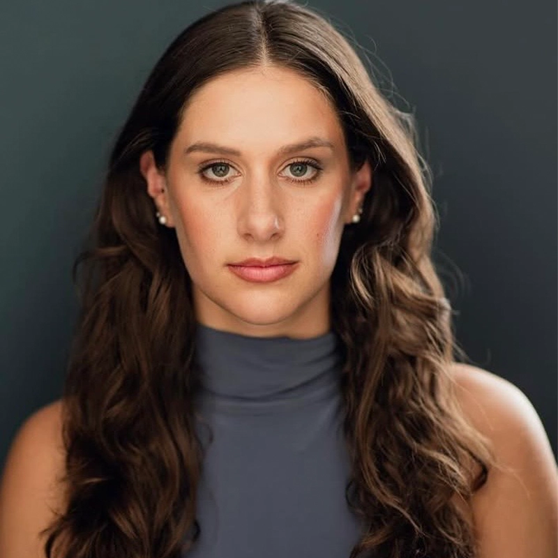 A woman with long wavy brown hair and fair skin looks directly at the camera with a neutral expression. She is wearing a sleeveless, high-necked gray top. The background is a plain, dark bluish-gray color.