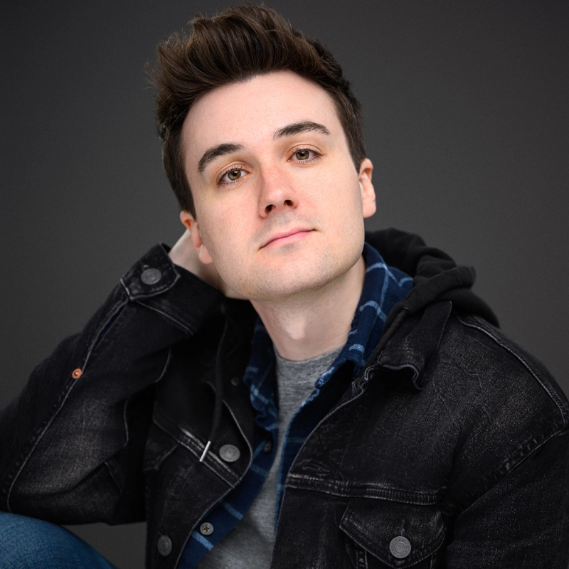 A young man with fair skin and brown hair styled up gazes softly at the camera. He wears a black denim jacket over a blue plaid shirt and gray t-shirt. One hand is behind his neck, and the background is solid dark gray.