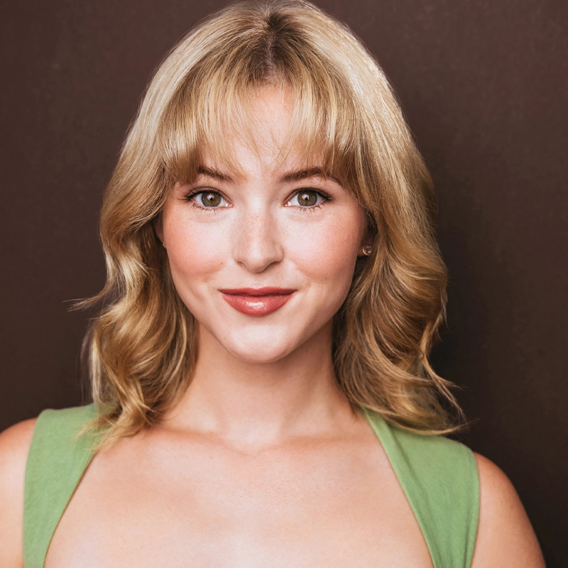 A young woman, Caroline Portner, with wavy blonde hair and bangs smiles at the camera. She is wearing a sleeveless green top and subtle makeup, with a brown background behind her. The image is well-lit, highlighting her clear complexion and expressive eyes.