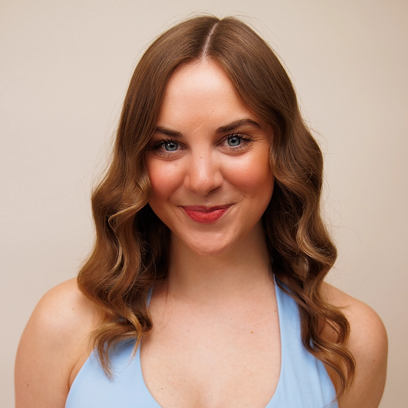A young woman, Quincy Lawson, with wavy light brown hair and blue eyes smiles at the camera. She is wearing a light blue sleeveless top against a soft beige background, her natural makeup highlighting rosy cheeks and pink lips.