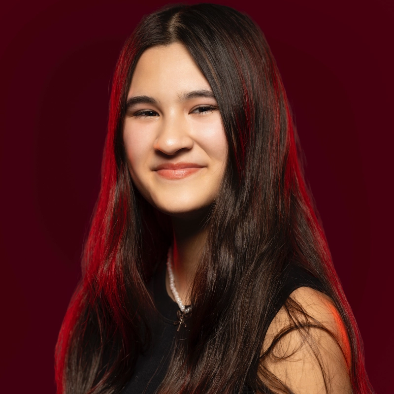 Lena Racelis, a young woman with long, dark brown hair and fair skin, smiles softly. She wears a sleeveless black top and a pearl necklace. The deep red background and red lighting highlight her hair and shoulders.