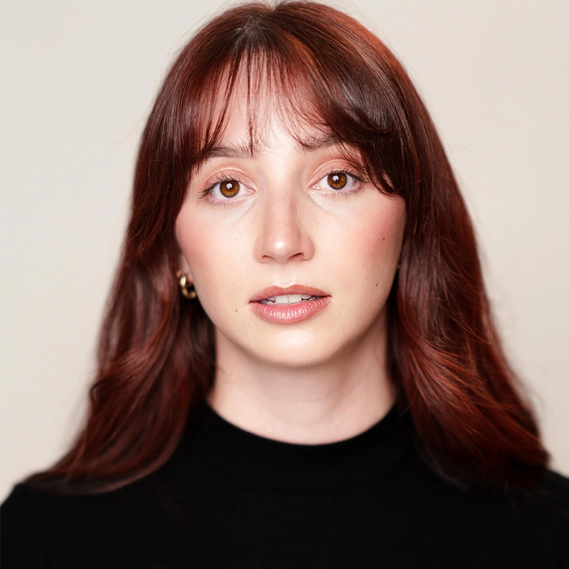 A young woman with fair skin, straight auburn hair with bangs, and brown eyes looks directly at the camera. She wears a black top and gold hoop earrings, embodying Quincy Lawson's timeless style against a plain, soft beige background.