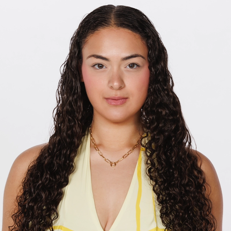 Renee Elkady, with long, dark, curly hair and medium skin tone, faces forward against a plain white background. She wears a light yellow halter top and a gold chain necklace. Her makeup is natural, with rosy cheeks and pink lips.