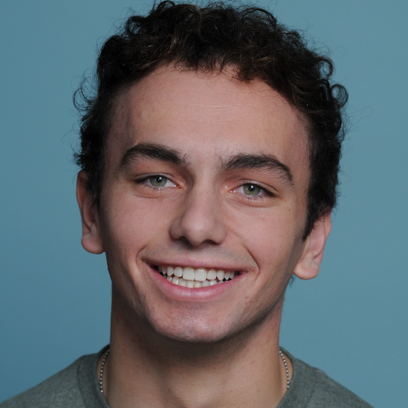 A young man with short, curly dark hair smiles broadly at the camera. Liam Restivo has light skin, green eyes, and wears a gray shirt. The background is a solid light blue. His expression is friendly and inviting.