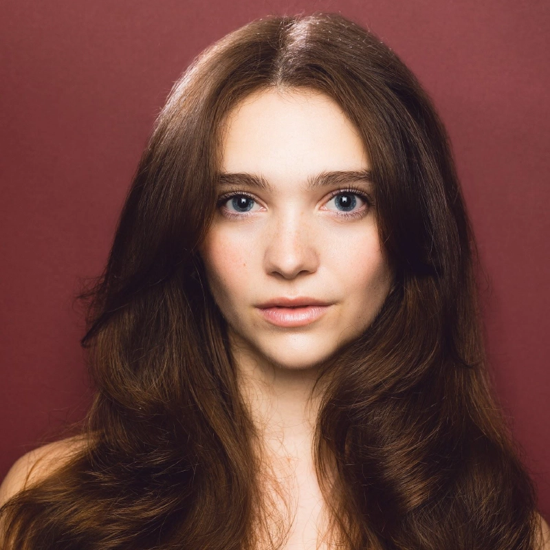 A young woman with long, wavy brown hair and fair skin looks directly at the camera. She has blue eyes and subtle makeup. Set against a solid maroon background, this soft and elegant portrait is by Josiah Jennings.