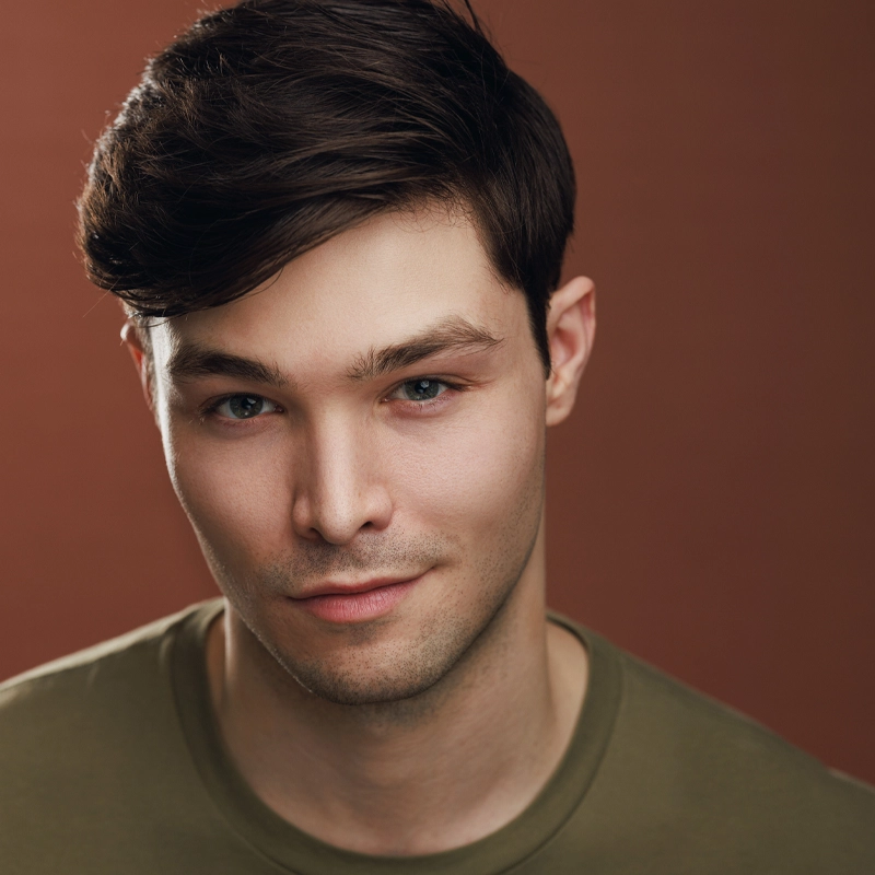 Alexander Rios, a young man with fair skin, short dark brown hair, and blue eyes, looks at the camera with a slight smile. He is wearing an olive green t-shirt and is posed in front of a plain, reddish-brown background.