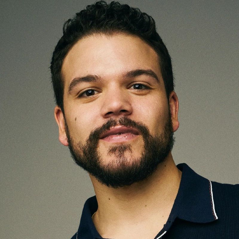 Josh Andrés Rivera, a man with short, curly dark hair and a trimmed beard, looks at the camera with a slight smile. He is wearing a navy collared shirt with white trim against a softly lit, neutral-toned background.