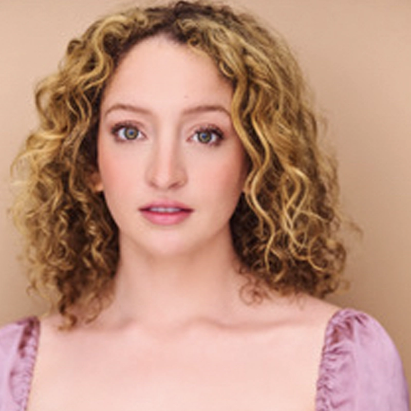 Catherine Roddey, a young woman with light skin and curly blonde hair, poses against a beige background. She has green eyes, subtle makeup, and wears a light purple square-neck top. Her expression is calm as she looks directly at the camera.