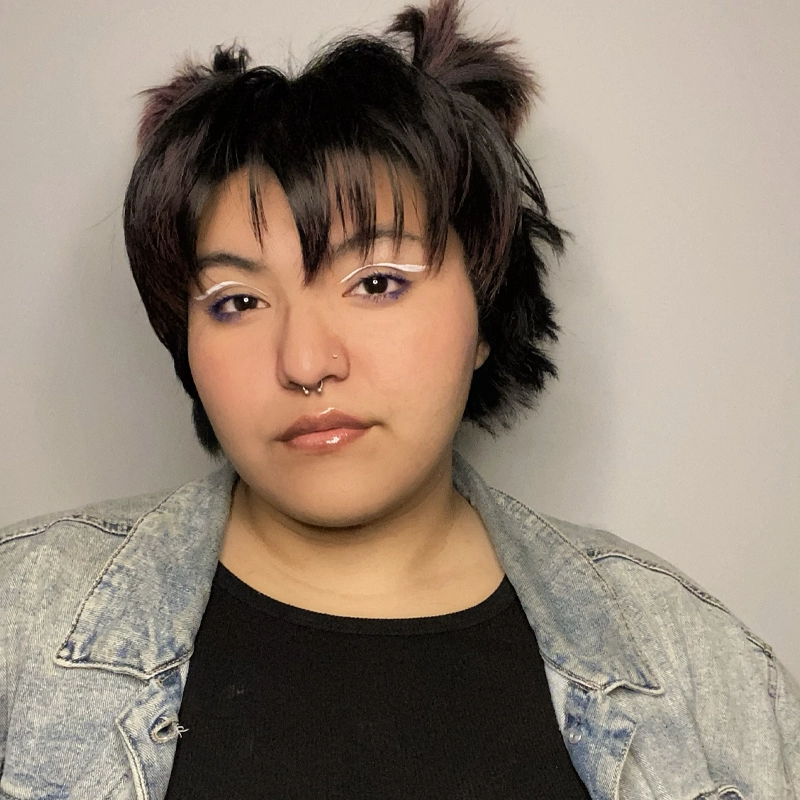 Person with short, dark, choppy hair styled with two small puffs, wearing bold white eyeliner, glossy lips, a nose ring, black top, and a light denim jacket stands against a plain light background—Angel Rodela looks calmly at the camera.